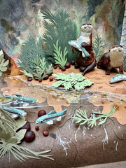 Diorama sculpture of 2 brown otters, one holds a fish in its hand and the other stands near a beached fish. They stand on a dirt shore with fish swimming nearby and cut greenery all around.