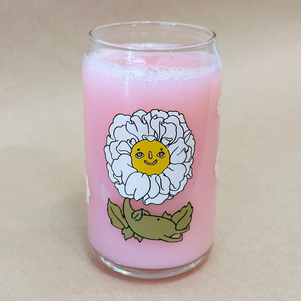 Glass cup with a flat base and slightly inward lip. Features a graphic of a cartoon style white camellia, sitting with a smiling face and a chubby green body made out of stems and leaves. Around the rest of the glass are white flowers.