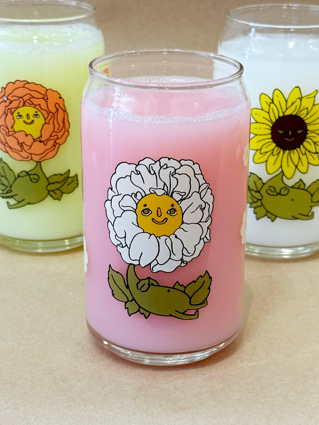 Glass cup with a flat base and slightly inward lip. Features a graphic of a cartoon style white camellia, sitting with a smiling face and a chubby green body made out of stems and leaves. Around the rest of the glass are white flowers.