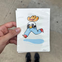 A hand holds an artwork featuring a cartoon character wearing a yellow-orange cowboy hat, a black-and-white striped shirt, blue pants, and red shoes, with a light blue splash underneath.