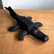 Whittled wooden sculpture of an alligator, painted all black and red eyes and a red mouth. Its underside is gray. Its body has a slightly blocky look to it, with its back being a row of cubes.