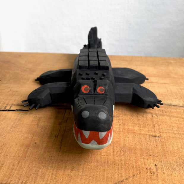 Whittled wooden sculpture of an alligator, painted all black and red eyes and a red mouth. Its underside is gray. Its body has a slightly blocky look to it, with its back being a row of cubes.