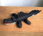 Whittled wooden sculpture of an alligator, painted all black and red eyes and a red mouth. Its underside is gray. Its body has a slightly blocky look to it, with its back being a row of cubes.