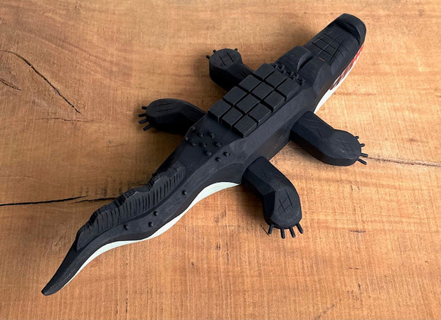 Whittled wooden sculpture of an alligator, painted all black and red eyes and a red mouth. Its underside is gray. Its body has a slightly blocky look to it, with its back being a row of cubes.