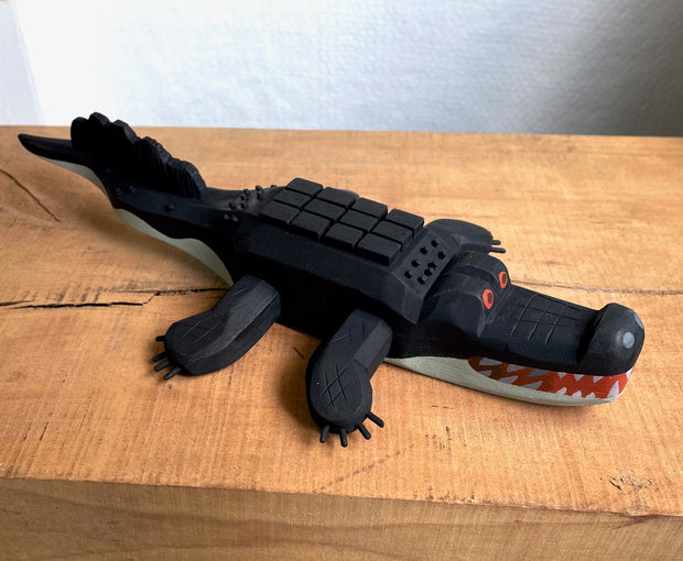 Whittled wooden sculpture of an alligator, painted all black and red eyes and a red mouth. Its underside is gray. Its body has a slightly blocky look to it, with its back being a row of cubes.