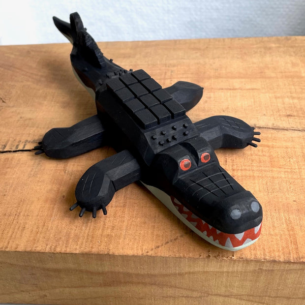 Whittled wooden sculpture of an alligator, painted all black and red eyes and a red mouth. Its underside is gray. Its body has a slightly blocky look to it, with its back being a row of cubes.