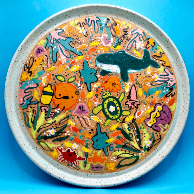 Round ceramic plate with many colorful drawings all along the main face of the plate with themes of underwater characters and creatures with seaweed and corals.