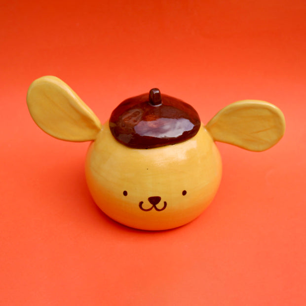 Round ceramic figure of a dog head made to look like Sanrio's Pompompurin with floppy ears going in different directions and his iconic brown beret.