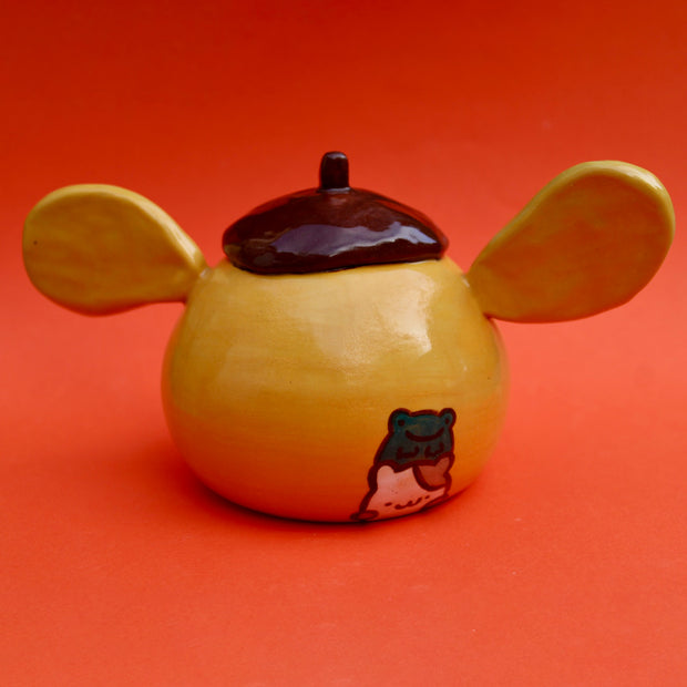 Round ceramic figure of a dog head made to look like Sanrio's Pompompurin with floppy ears going in different directions. Backside has a drawing of a cat and a frog stacked atop one another.