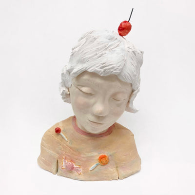 Ceramic sculpture of a bust of a young girl. She has frosting like hair and a cherry atop her head. She looks down with a closed eye, sleepy expression and wears a t-shirt with pieces of candy stuck to it.