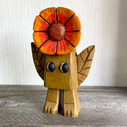 Small whittled wooden sculpture of a blocky brown creature with no facial features except for cute eyes pointed to the side. 2 leaves come out the side of its body and atop its head is a bright orange and red flower.