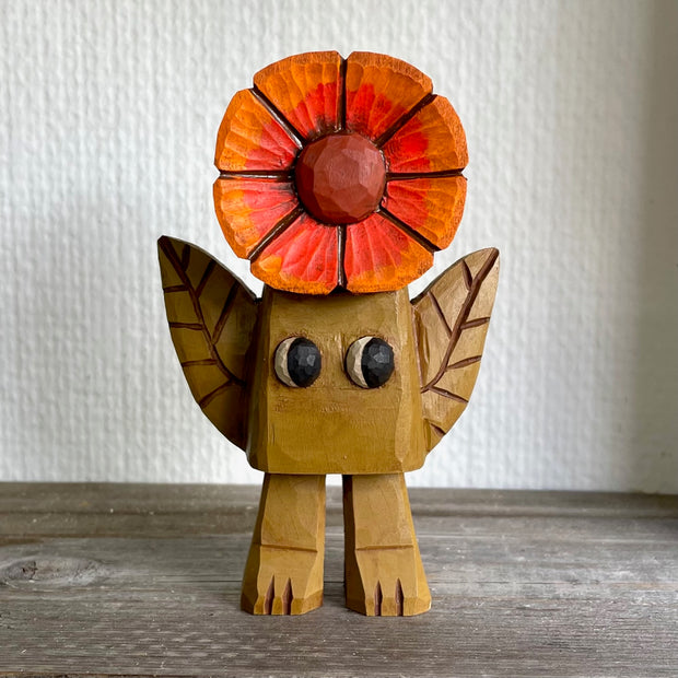 Small whittled wooden sculpture of a blocky brown creature with no facial features except for cute eyes pointed to the side. 2 leaves come out the side of its body and atop its head is a bright orange and red flower.