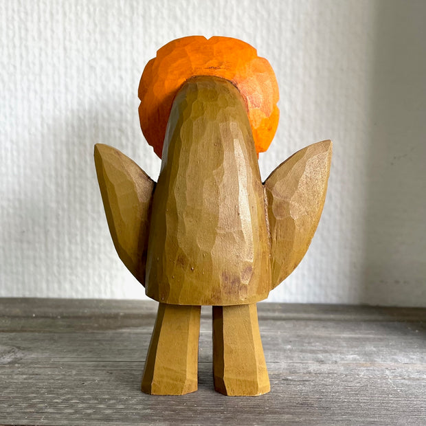 Small whittled wooden sculpture of a blocky brown creature with no facial features except for cute eyes pointed to the side. 2 leaves come out the side of its body and atop its head is a bright orange and red flower.