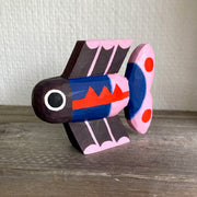 Brightly painted whittled wooden sculpture of a fish colored black with blue and pink body and tail and red accent coloring. It has a set of eyes, one on each side of its head.