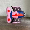 Brightly painted whittled wooden sculpture of a fish colored black with blue and pink body and tail and red accent coloring. It has a set of eyes, one on each side of its head.