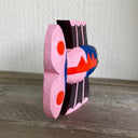 Brightly painted whittled wooden sculpture of a fish colored black with blue and pink body and tail and red accent coloring. It has a set of eyes, one on each side of its head.