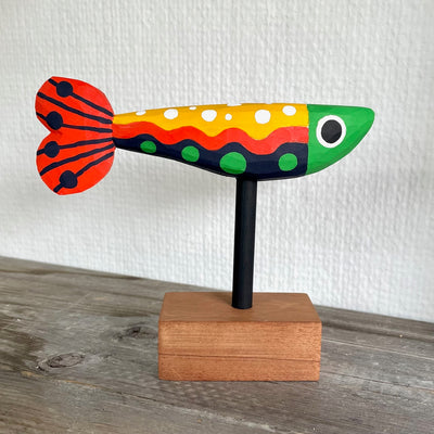 Colorfully painted whittled wooden fish, red green yellow and black. It has a polka dot pattern and one large eye on each side of its head. The fish is mounted on a rod attached to a small wooden block.