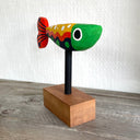 Colorfully painted whittled wooden fish, red green yellow and black. It has a polka dot pattern and one large eye on each side of its head. The fish is mounted on a rod attached to a small wooden block.