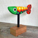 Colorfully painted whittled wooden fish, red green yellow and black. It has a polka dot pattern and one large eye on each side of its head. The fish is mounted on a rod attached to a small wooden block.