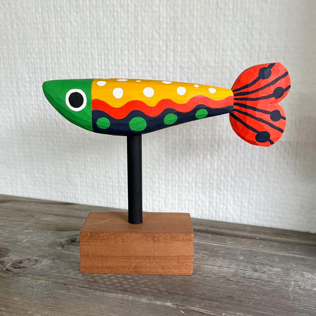 Colorfully painted whittled wooden fish, red green yellow and black. It has a polka dot pattern and one large eye on each side of its head. The fish is mounted on a rod attached to a small wooden block.