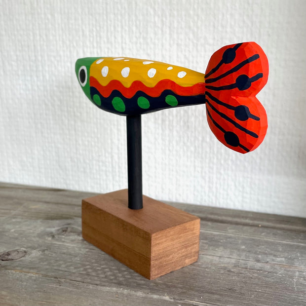Colorfully painted whittled wooden fish, red green yellow and black. It has a polka dot pattern and one large eye on each side of its head. The fish is mounted on a rod attached to a small wooden block.