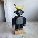 Assembled wooden sculpture of a smiling cartoon gorilla with a thin neck and thick arms and legs. Balancing atop its head is a banana.