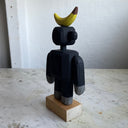 Assembled wooden sculpture of a smiling cartoon gorilla with a thin neck and thick arms and legs. Balancing atop its head is a banana.