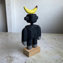 Assembled wooden sculpture of a smiling cartoon gorilla with a thin neck and thick arms and legs. Balancing atop its head is a banana.