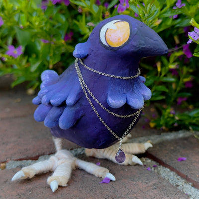 Sculpture of a purple bird with large yellow cartoon eyes and large clawed feet. It has a silver necklace with a purple gem wrapped around its neck.