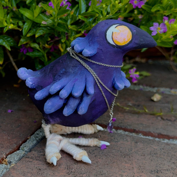 Sculpture of a purple bird with large yellow cartoon eyes and large clawed feet. It has a silver necklace with a purple gem wrapped around its neck.