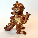 Ceramic sculpture of Garfield, entirely a muddy brown color. He is modeled like Godzilla with a large kaiju tail and holds a decapitated creature in his hand