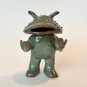 Ceramic sculpture of a catfish like creature, standing with a humanoid body. It has an open mouth and holds its hands slightly up.