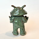 Ceramic sculpture of a catfish like creature, standing with a humanoid body. It has an open mouth and holds its hands slightly up.