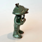 Ceramic sculpture of a catfish like creature, standing with a humanoid body. It has an open mouth and holds its hands slightly up.