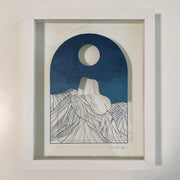 Delicate paper cutting made out of blue dyed paper of a woman's nude body sitting atop of a mountain range. She has no head and is turned away so only her back and butt are showing. A full moon is in the night sky.