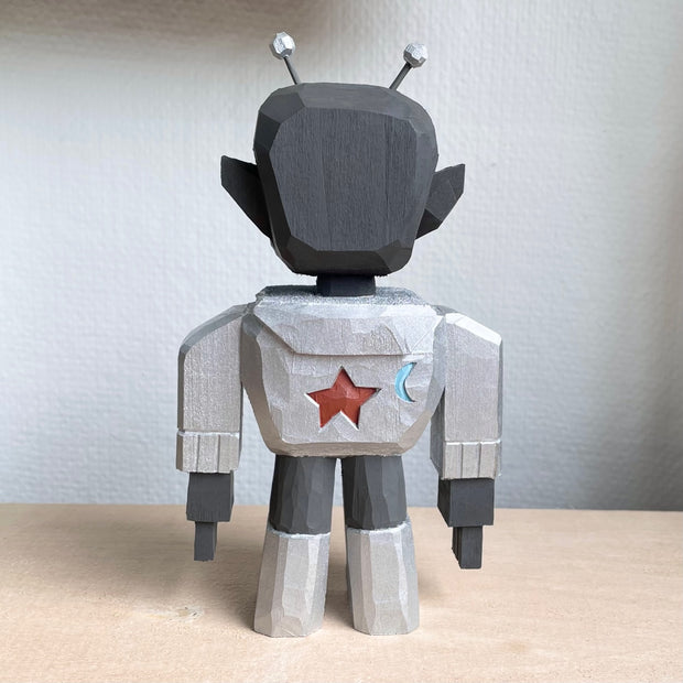 Whittled wooden sculpture of a grey alien, wearing silver futuristic space gear and funky comma shaped red sunglasses. It has a set of silver antennae and grits its teeth.