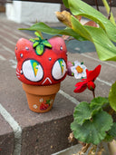 Sculpture in a small planter pot of a large cartoon cat head, made to look like a strawberry with large green and blue eyes. It has no limbs but a small white flower blooms from its tail.
