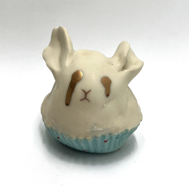 Cream colored sculpture of a bunny shaped cupcake, in a blue liner. It has dripping gold eyes and a simple face.