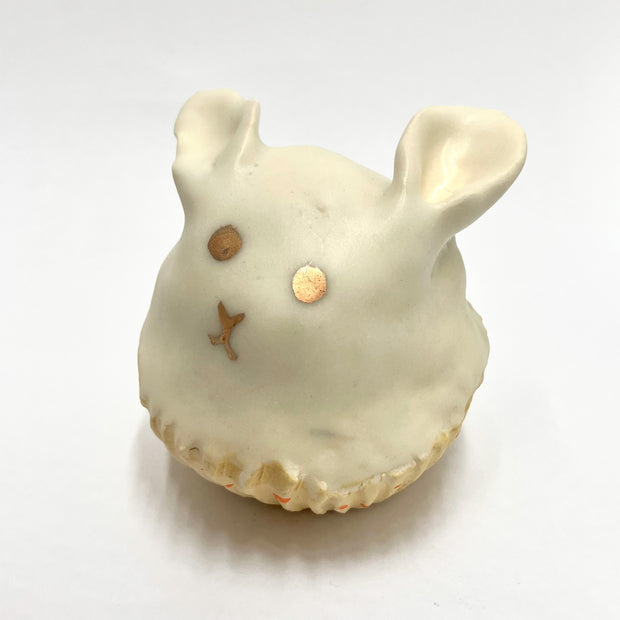 Cream colored sculpture of a bunny shaped cupcake, in a liner. It has gold eyes and a simple face.