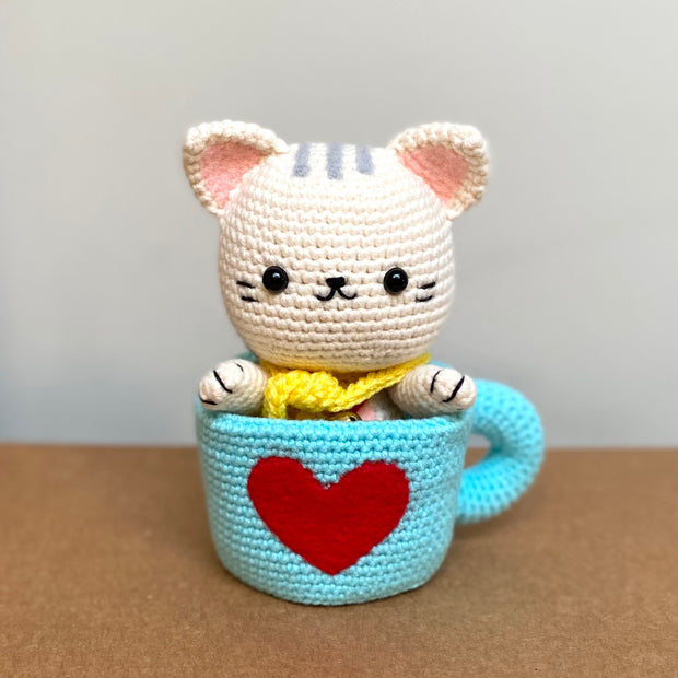 Crochet sculpture of a white cat in a cute, chibi style wearing a yellow scarf and sitting in a blue mug with a red heart on the exterior.