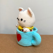Crochet sculpture of a white cat in a cute, chibi style wearing a yellow scarf and sitting in a blue mug with a red heart on the exterior.