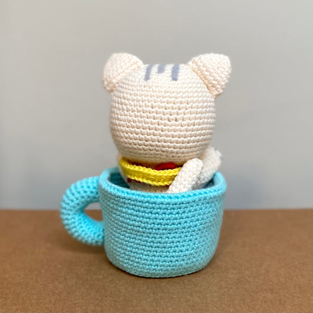 Crochet sculpture of a white cat in a cute, chibi style wearing a yellow scarf and sitting in a blue mug with a red heart on the exterior.