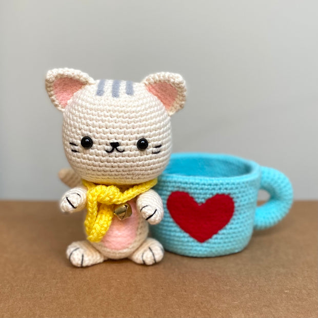 Crochet sculpture of a white cat in a cute, chibi style wearing a yellow scarf and sitting next to a blue mug with a red heart on the exterior.