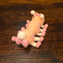 Felted sculpture of a pastel orange caterpillar with a small smiling face and 10 legs, all spread out flat so it is laying instead of standing. It wears pink shoes on all its feet and has a matching pink bow.