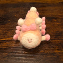Felted sculpture of a pastel orange caterpillar with a small smiling face and 10 legs, all spread out flat so it is laying instead of standing. It wears pink shoes on all its feet and has a matching pink bow.