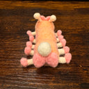 Felted sculpture of a pastel orange caterpillar with a small smiling face and 10 legs, all spread out flat so it is laying instead of standing. It wears pink shoes on all its feet and has a matching pink bow.