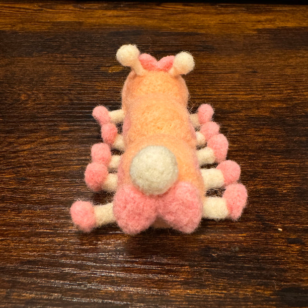 Felted sculpture of a pastel orange caterpillar with a small smiling face and 10 legs, all spread out flat so it is laying instead of standing. It wears pink shoes on all its feet and has a matching pink bow.