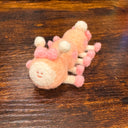 Felted sculpture of a pastel orange caterpillar with a small smiling face and 10 legs, all spread out flat so it is laying instead of standing. It wears pink shoes on all its feet and has a matching pink bow.