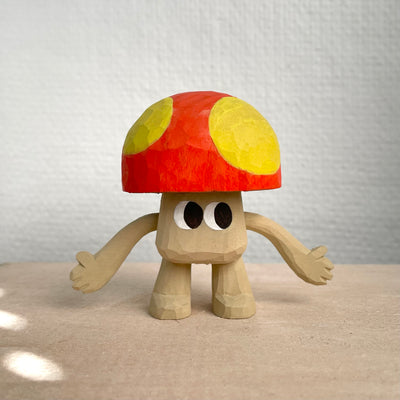 Whittled wooden sculpture of a mushroom with arms and large cartoon eyes and no other facial features. It's body is the exposed natural wood color and its cap is red with large yellow spots.