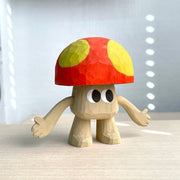 Whittled wooden sculpture of a mushroom with arms and large cartoon eyes and no other facial features. It's body is the exposed natural wood color and its cap is red with large yellow spots.